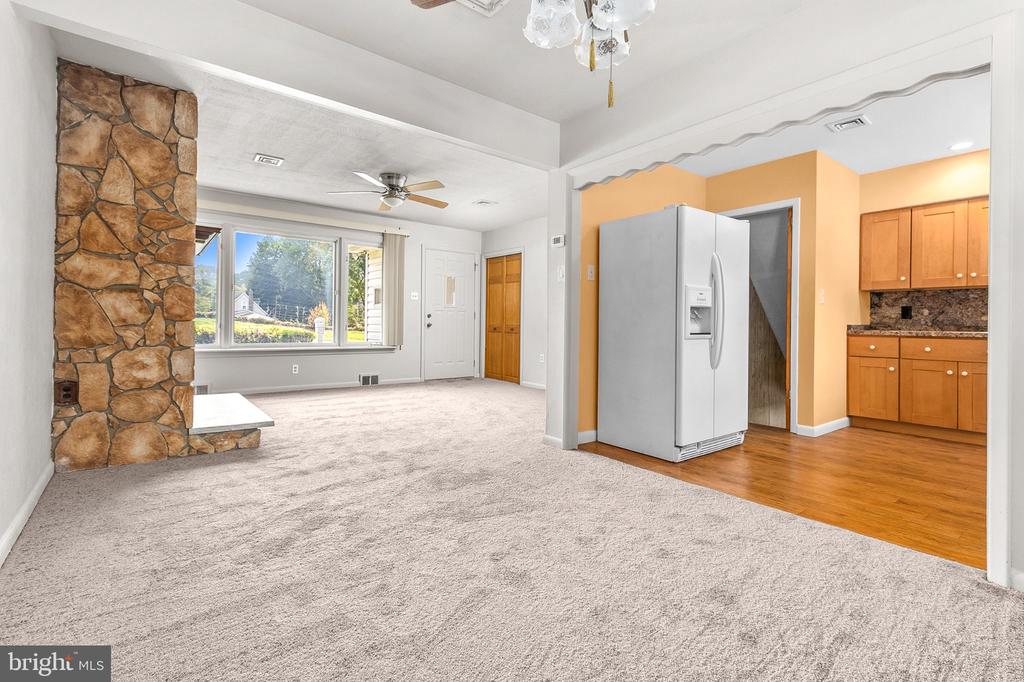 Living room with a stone fireplace, wide window, carpeted floor, and adjoining kitchen with wood cabinets.
