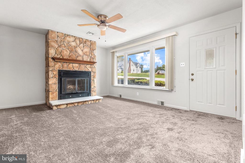 Vacant living room with a stone fireplace, ceiling fan, and large front window.