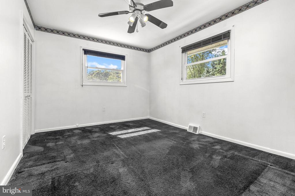 Empty bedroom with dark carpet, two windows, louvered closet doors, and a ceiling fan.