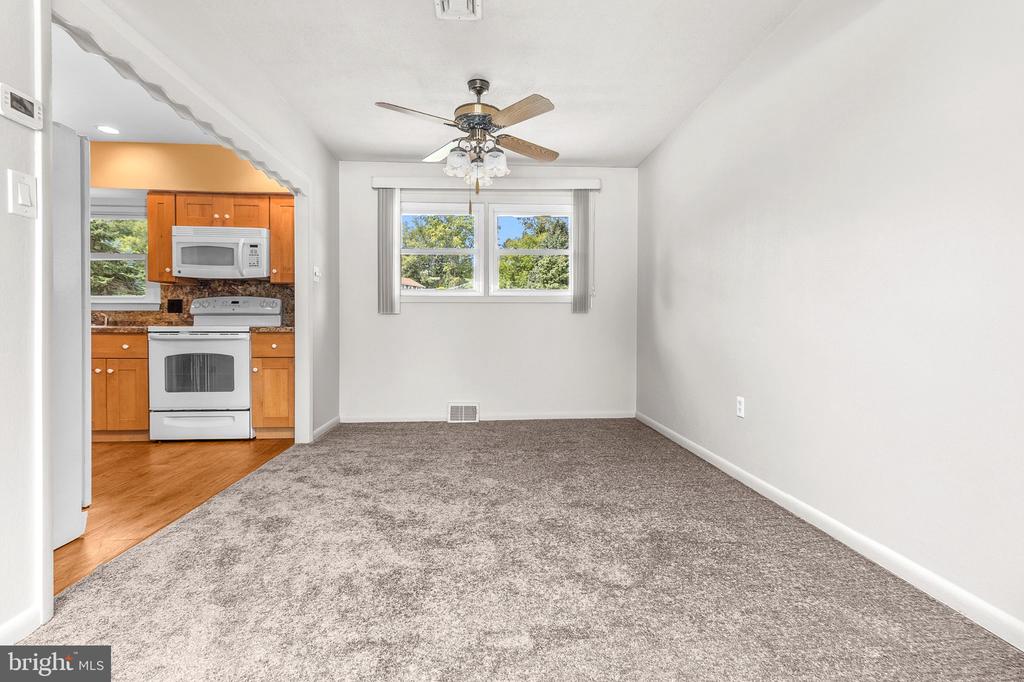 Empty carpeted living area opening to a kitchen with warm wood cabinets and white appliances.