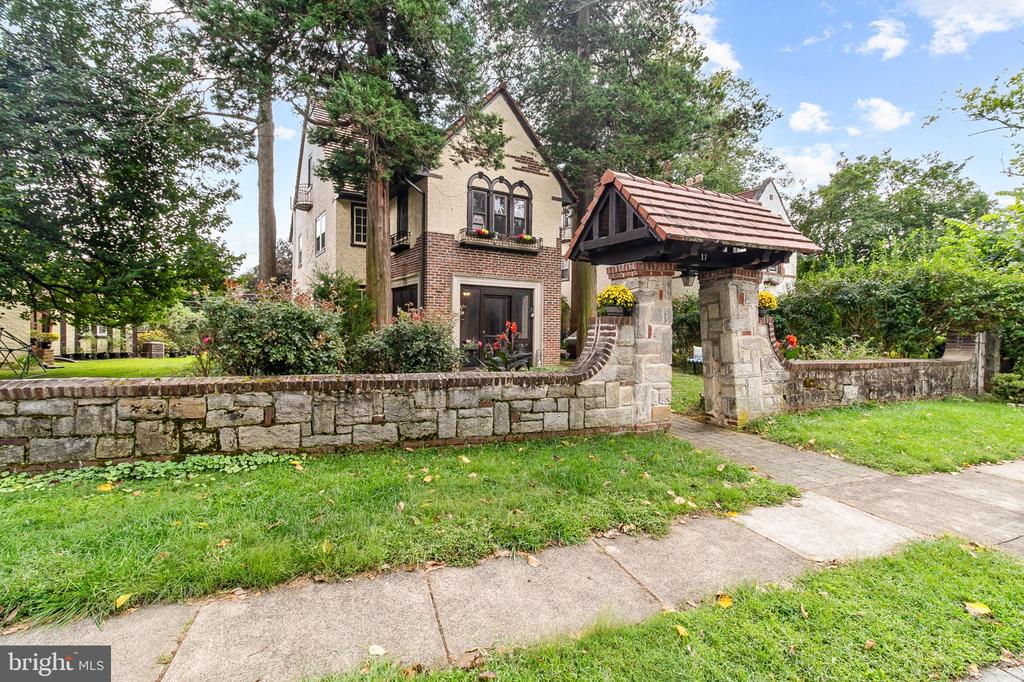 Tudor-style brick house behind a stone wall, with a tiled-roof entrance and mature trees.