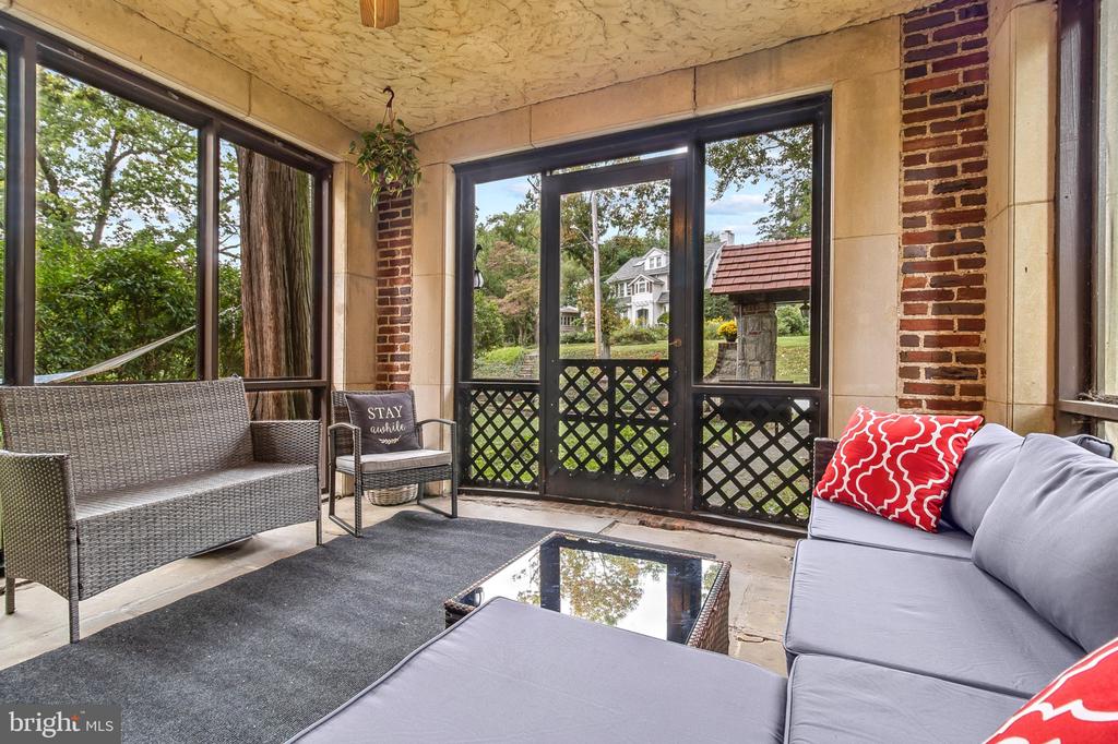 Enclosed porch with gray seating, a glass-top coffee table, and windows overlooking a leafy yard.