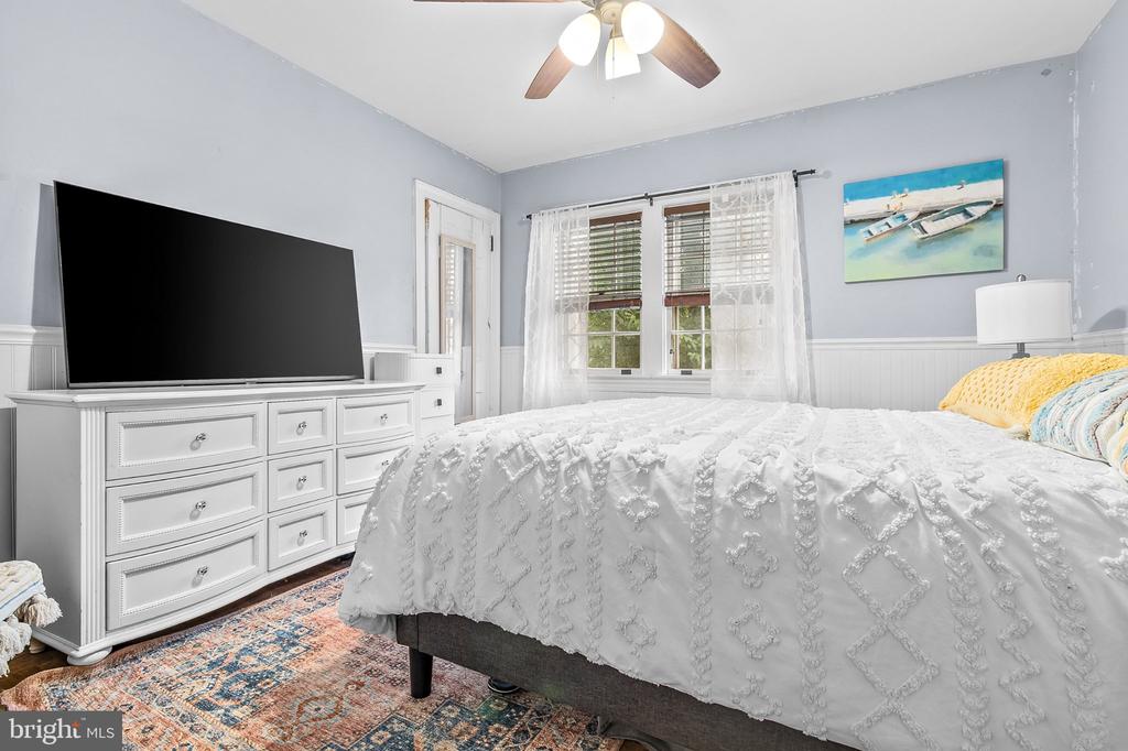 Bedroom with a white quilted bed, dresser-mounted television, and sheer-curtained windows.