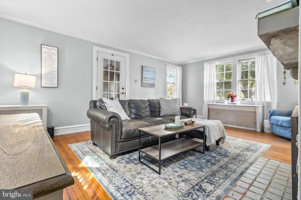 Living room with a dark leather sofa, patterned rug, hardwood floors, and bright curtained windows.