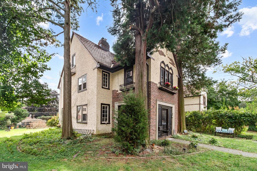 Stucco and brick home with small balconies, a steep tiled roof, and mature trees in the garden.