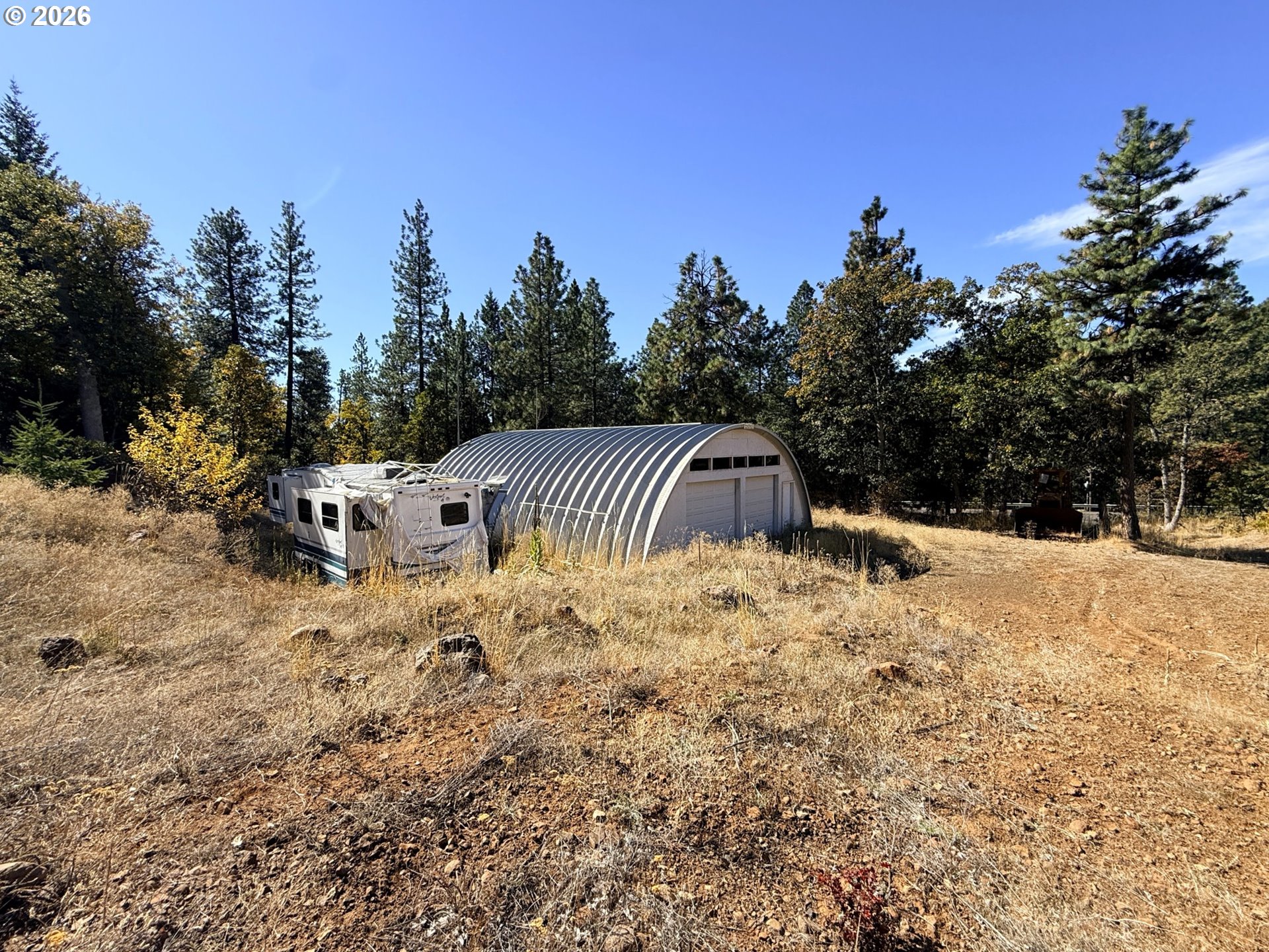 Arched metal garage and white RV in a dry, wooded lot.