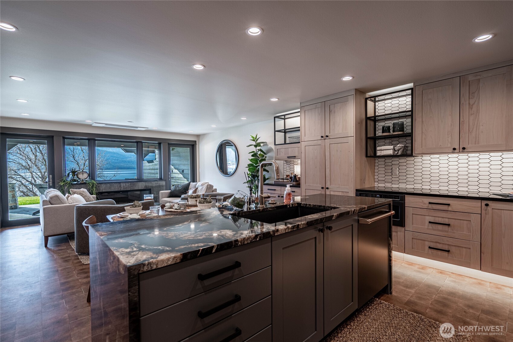 Open-plan kitchen with a large dark stone island, pale wood cabinets, and water and mountains beyond the windows.