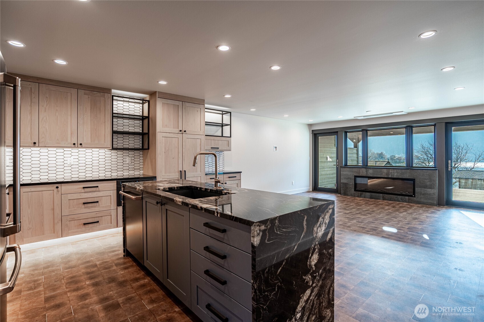 Open-plan kitchen with a large veined stone island, opening onto a living area with a fireplace and wide windows.