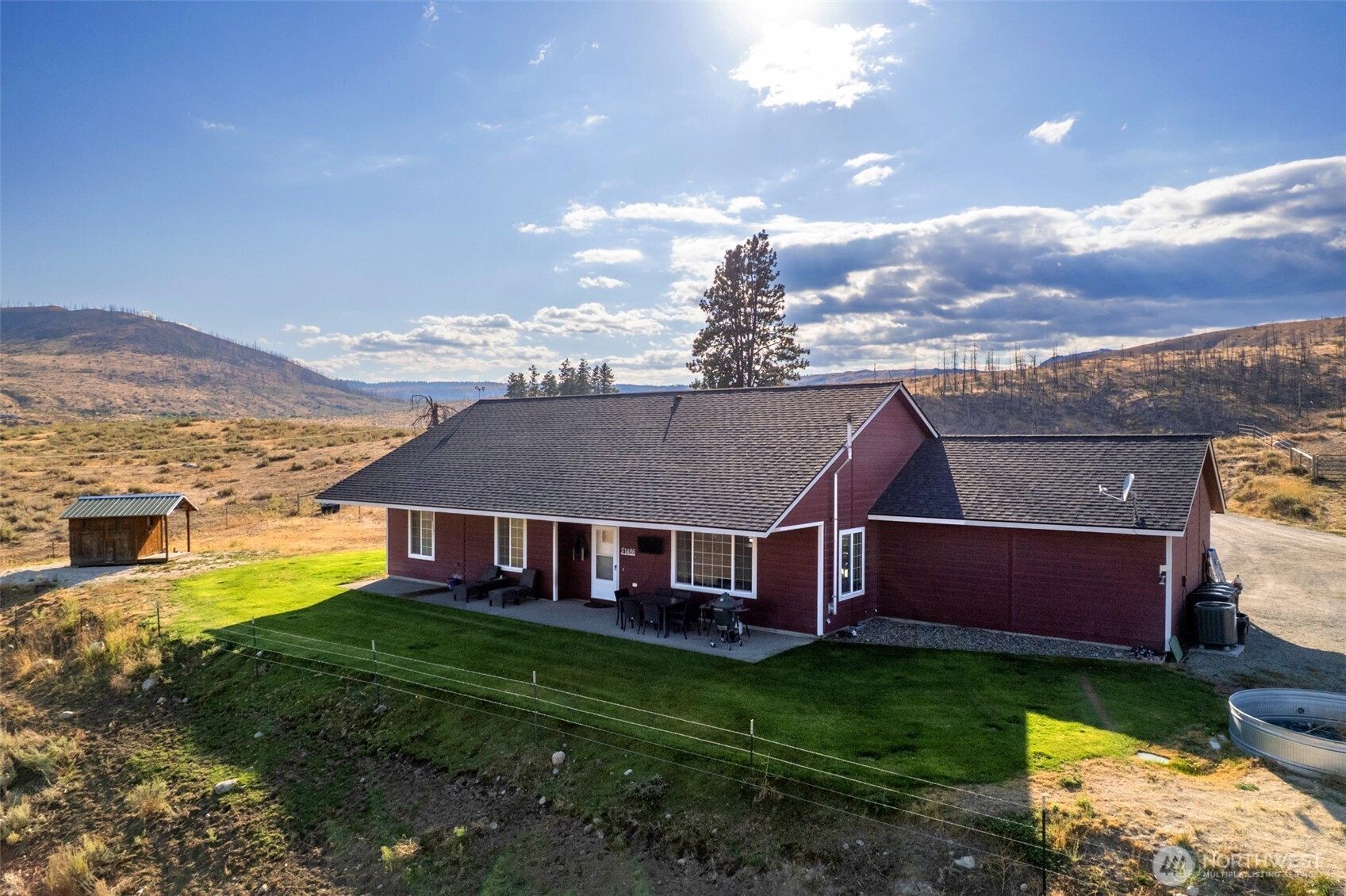 Red single-story rural home with a green lawn, patio furniture, and rolling hills beyond.