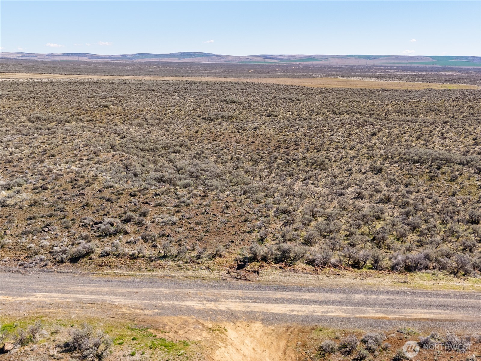 Vacant shrub-covered lot beside a gravel road, with open hills in the distance.