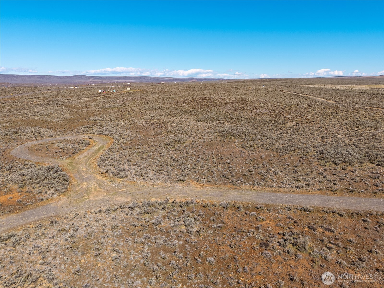Aerial view of open scrubland with a curving dirt track and distant buildings.