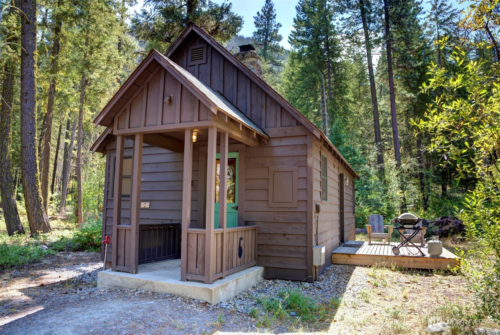 Small brown cabin with a turquoise door, covered porch, and deck among tall trees.