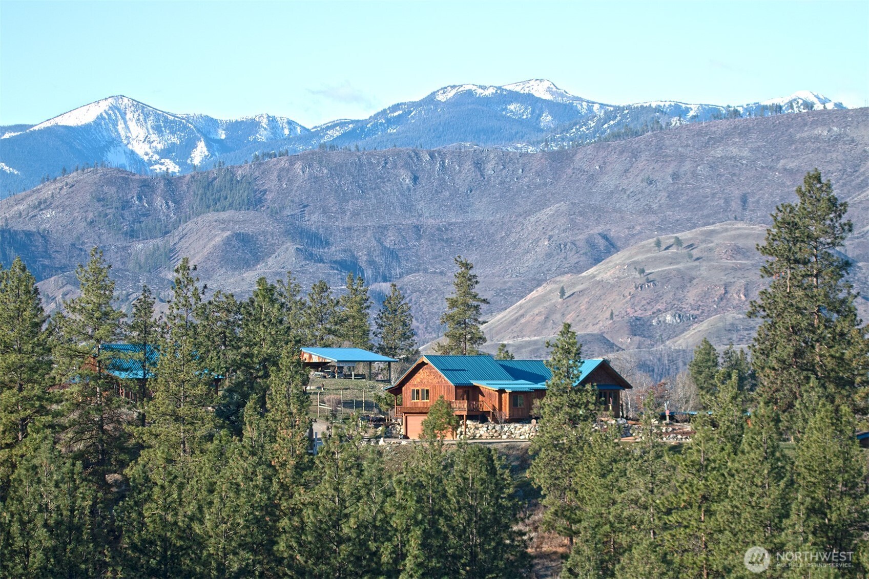 Wood-sided home with blue metal roofs among pine trees, backed by snow-capped mountains.