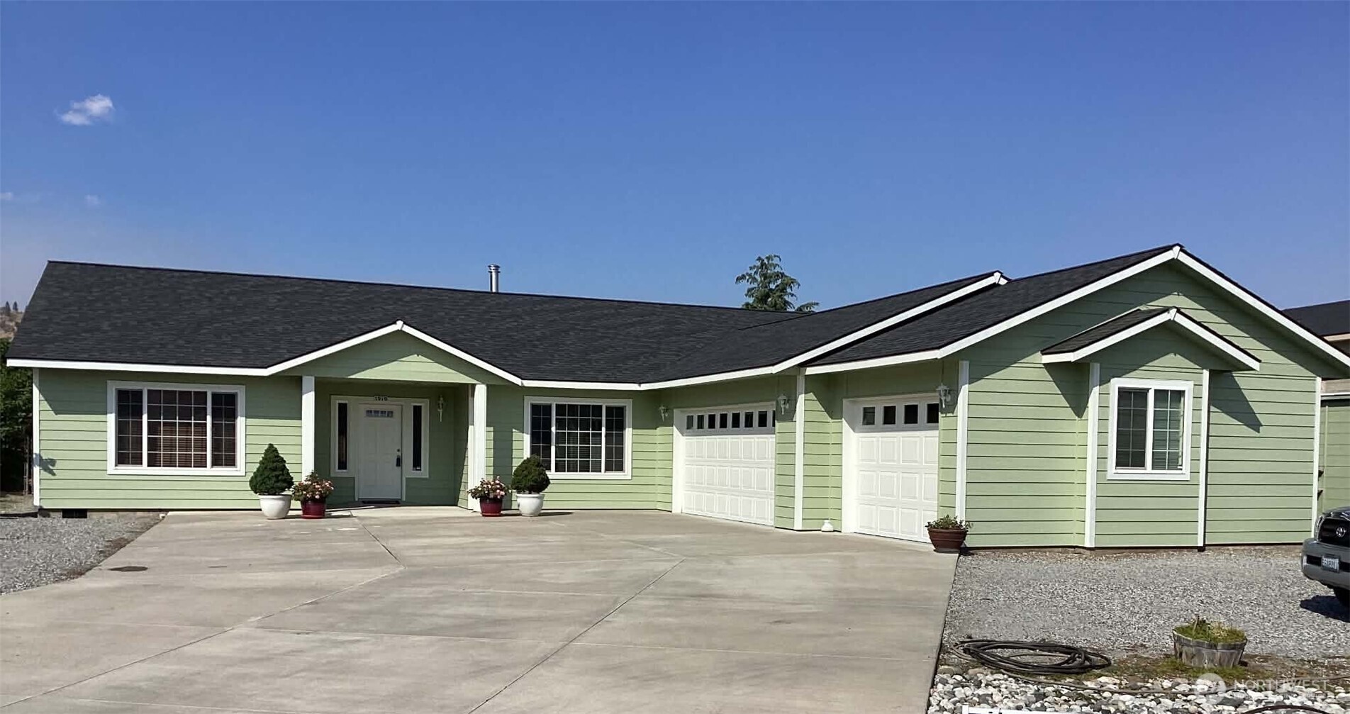 Light green single-story home with two garage doors, a broad driveway, and potted plants by the entrance.