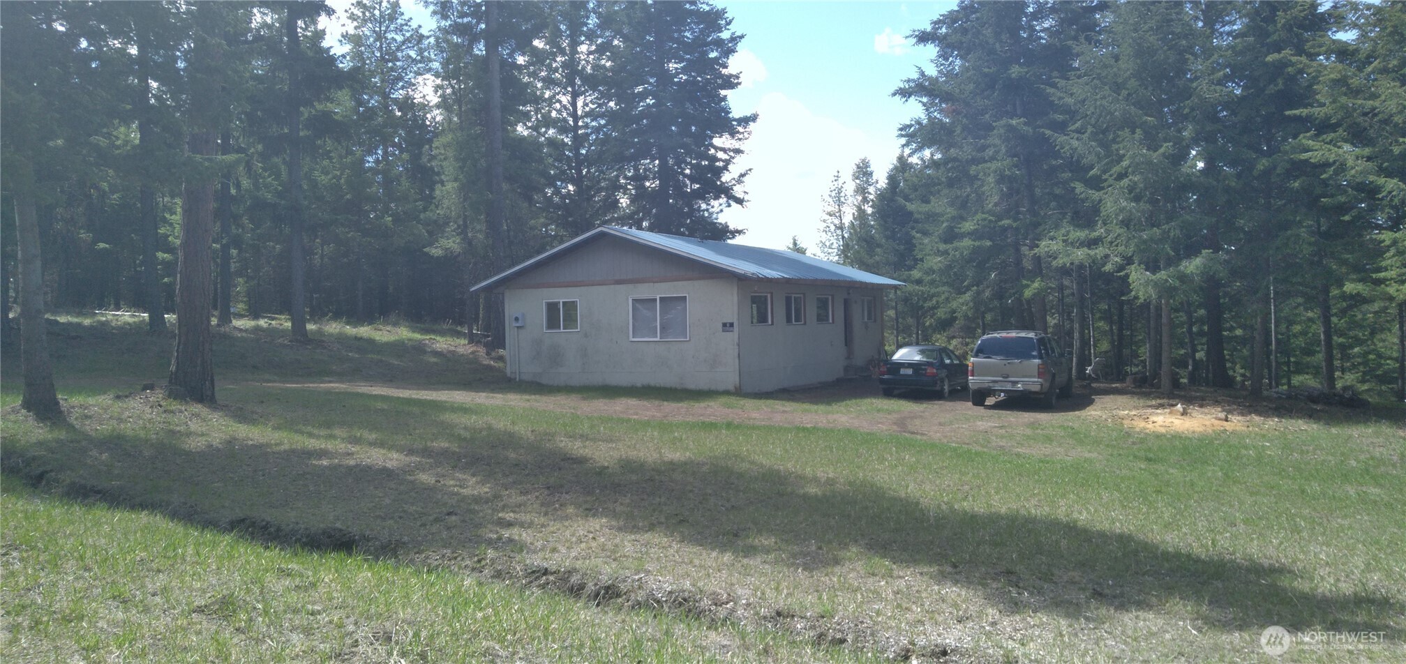 Single-story home in a wooded clearing, with a metal roof and two vehicles parked beside it.