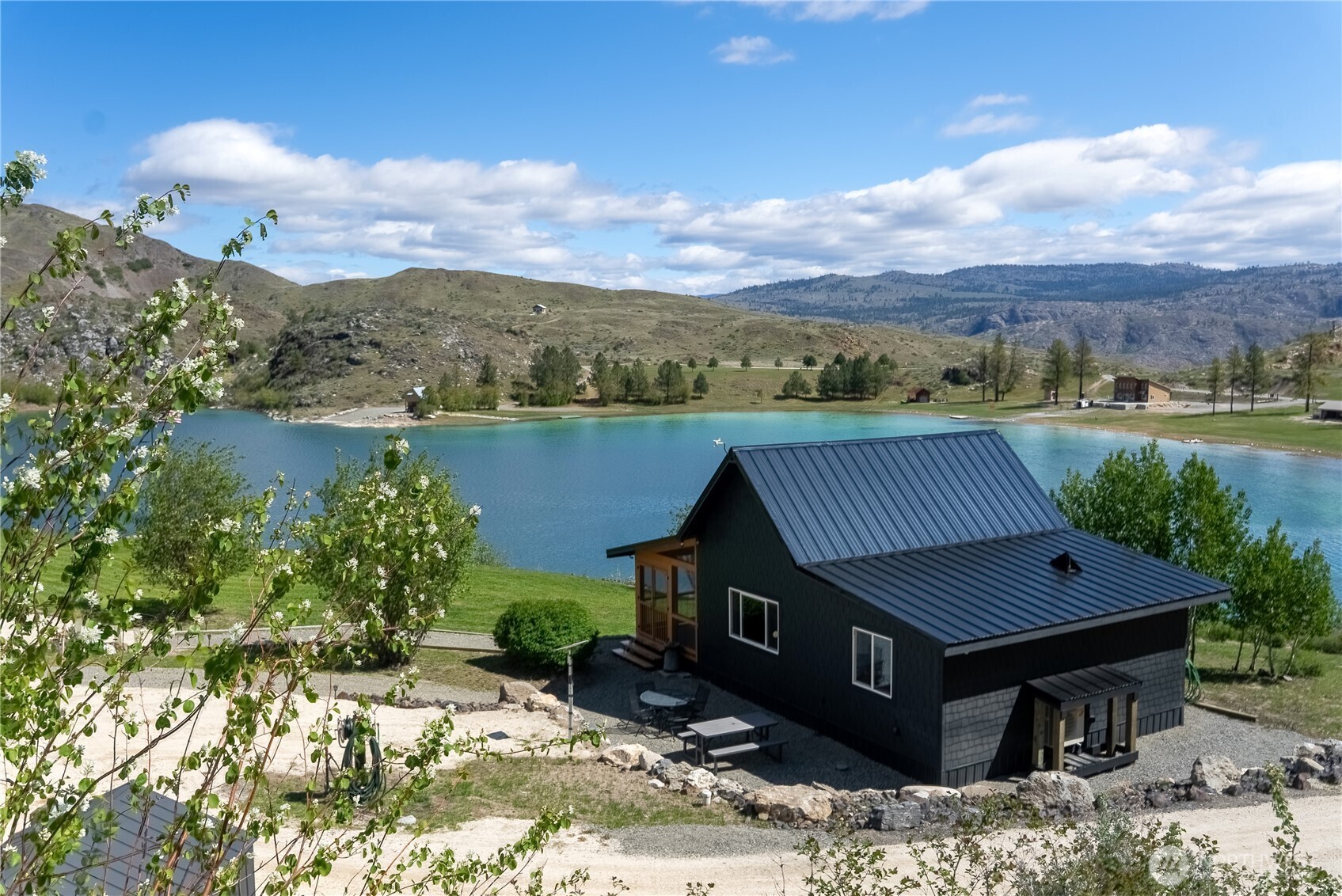 Dark metal-roofed lakeside home overlooking turquoise water, rolling hills, and distant mountains.