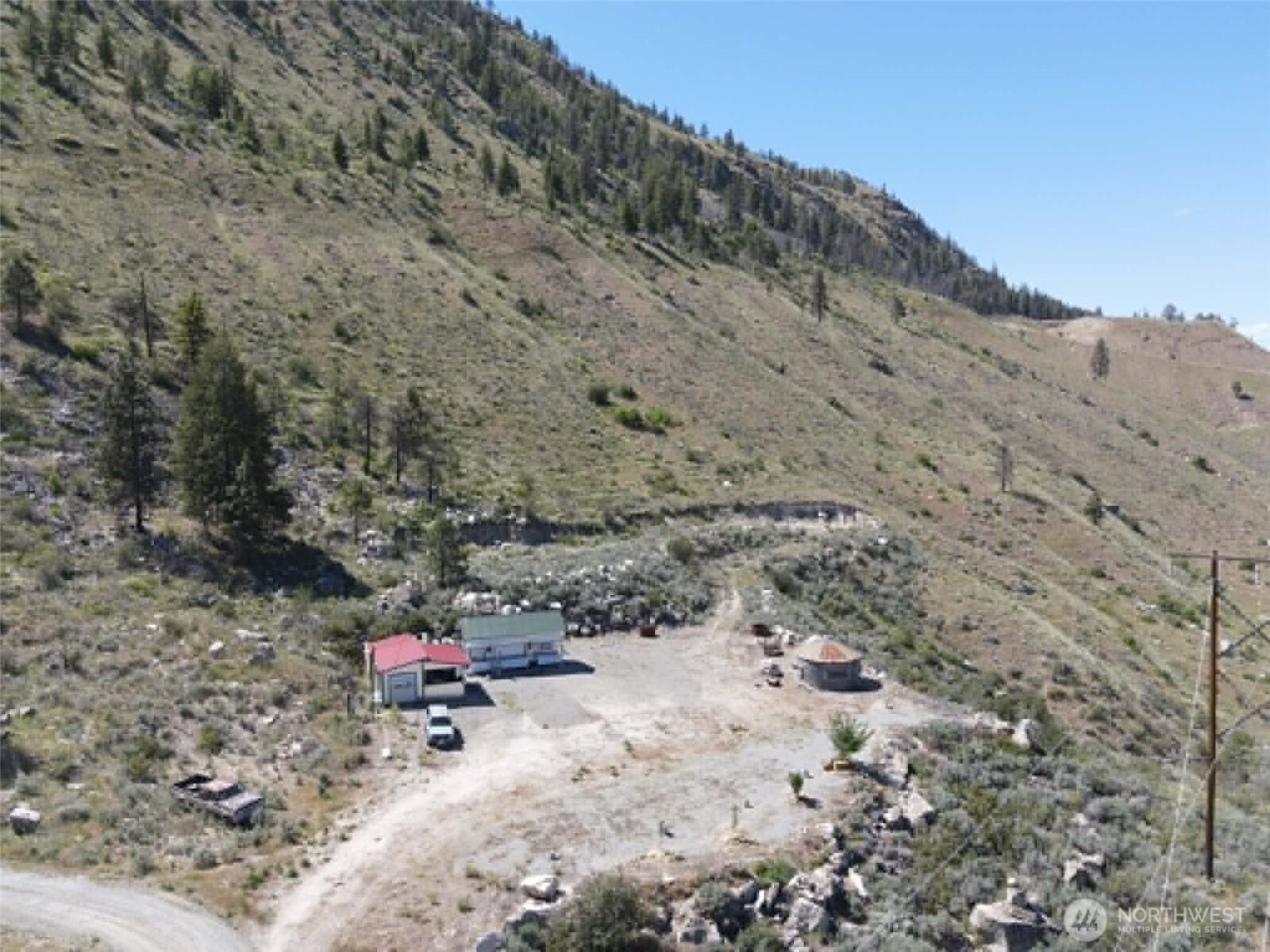 Rural hillside property with a gravel clearing, two small buildings, and sparse trees.