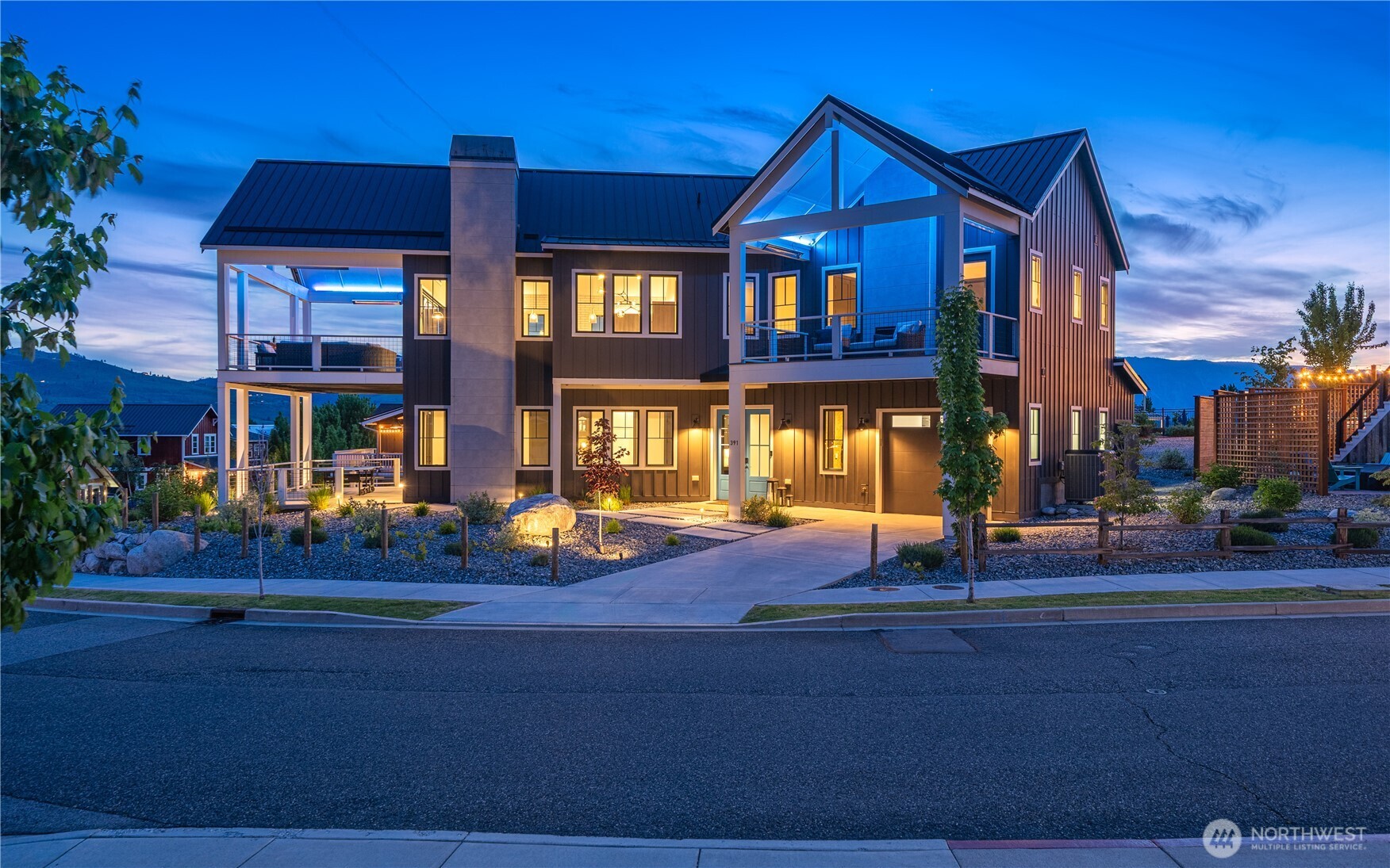 Modern two-story home at dusk with a glass-fronted gable, covered balconies, and illuminated landscaping.