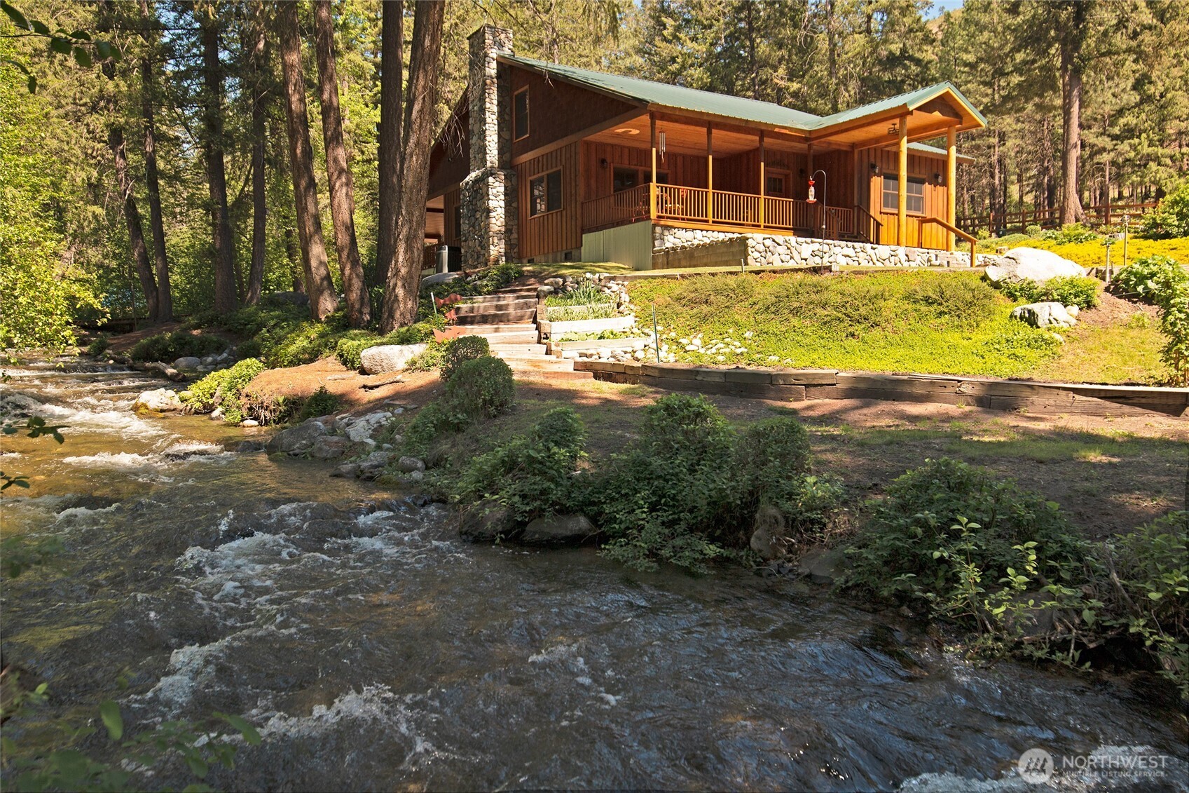 Wood-sided cabin with a stone chimney and covered porch beside a tree-lined creek.