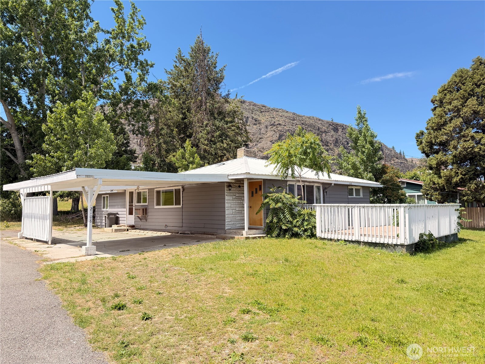 Single-story gray home with a white carport and raised deck, backed by trees and a rocky hillside.