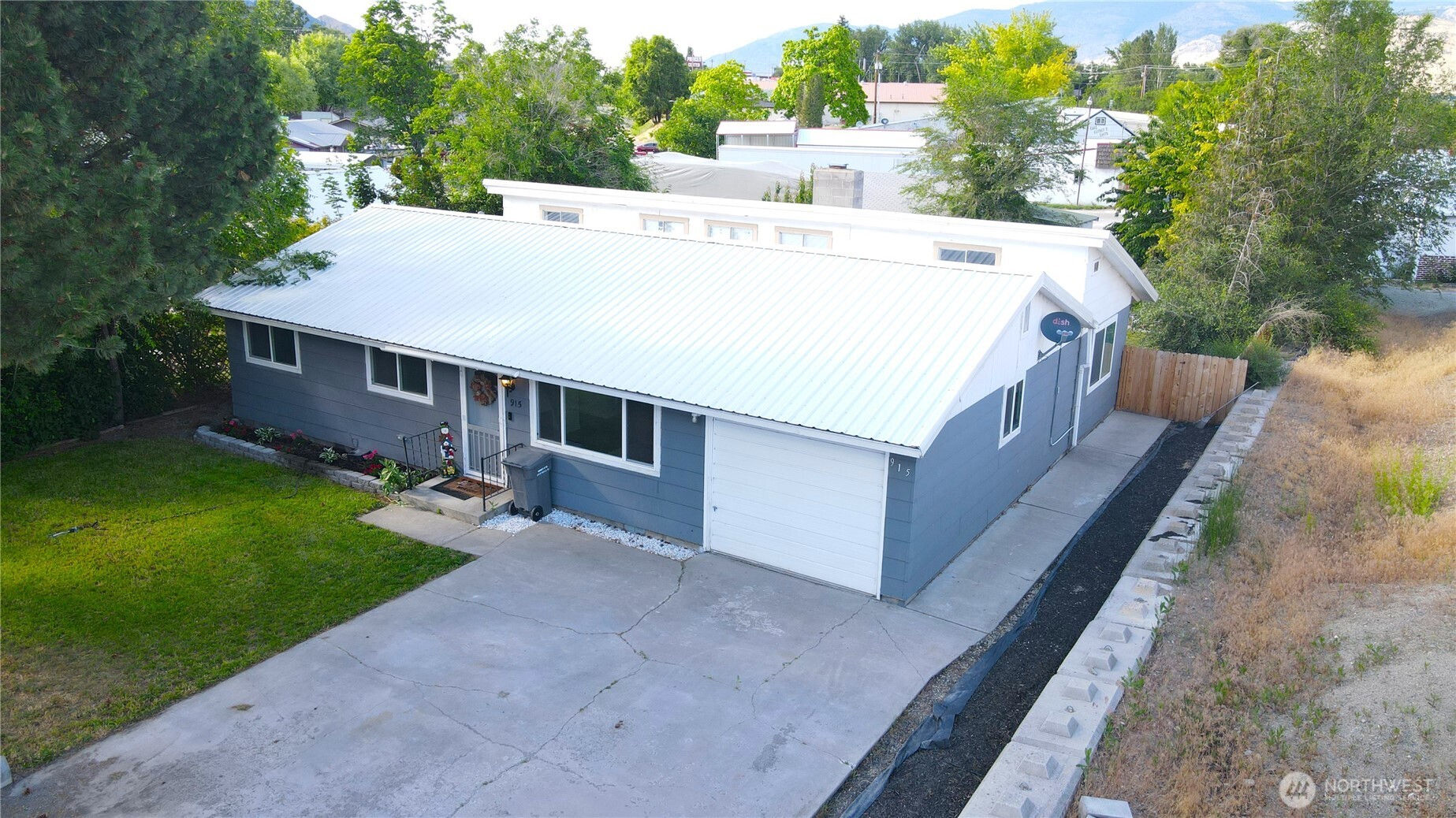 Blue-gray single-story house with a white metal roof, attached garage, and broad concrete driveway.