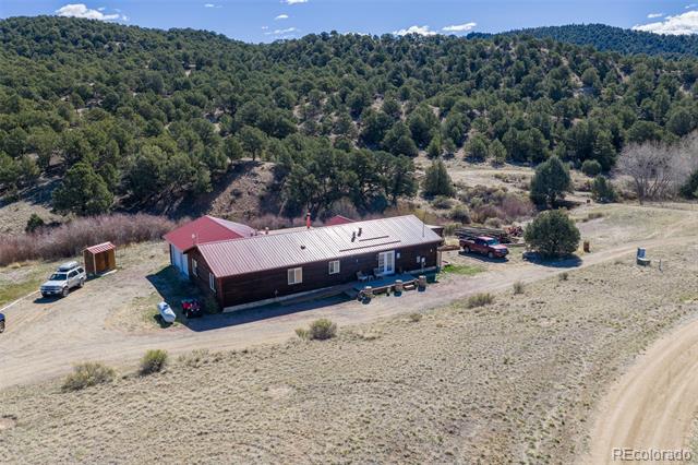 Aerial view of a long, metal-roofed property in a clearing, with wooded hills behind it.