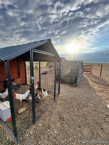 Chicken coop and fenced run with chickens, gravel yard, and open land at sunset.
