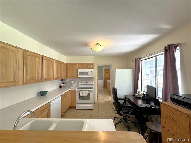 Kitchen with light wood cabinets and white appliances, opening onto a desk beside a large window.