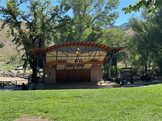 Covered outdoor stage with a curved wooden roof on a grassy lawn beneath mature trees.