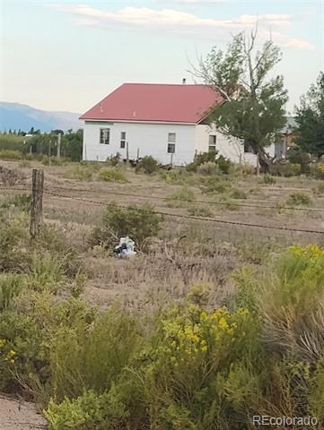 White rural house with a red metal roof behind a wire fence, with scrubland and distant mountains.