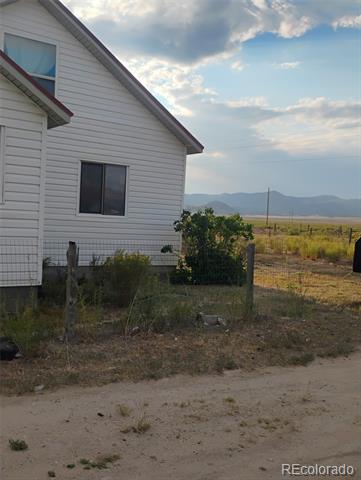 Light-sided house beside a dirt road, with a fenced yard and distant mountains.