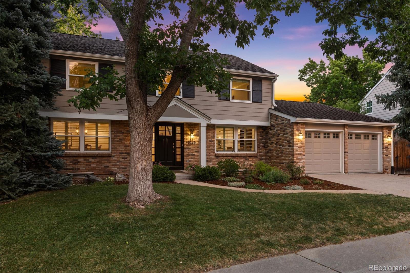 Two-story brick-and-siding home with a double garage and large front-yard tree at dusk.