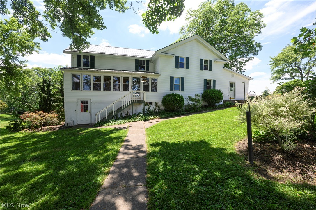 White two-story house with dark shutters, a long enclosed porch, and a walkway through a leafy lawn.