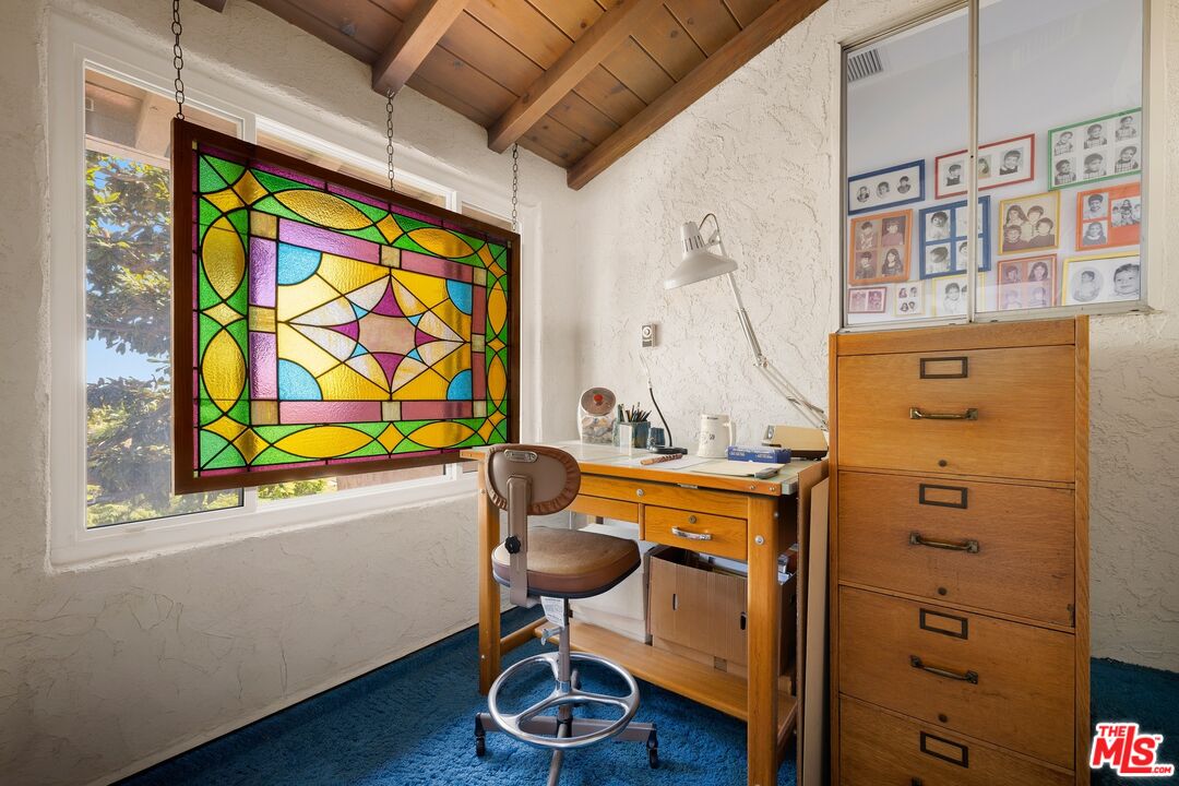 Home office with a colorful hanging stained-glass panel, wooden desk, and vaulted wood ceiling.