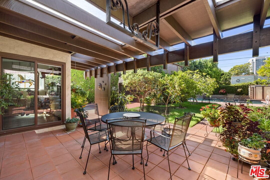Furnished tiled patio beneath a timber pergola, with a garden and swimming pool beyond.
