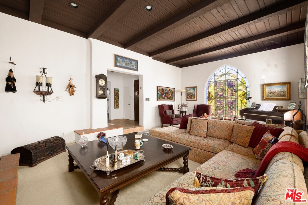 Furnished living room with a patterned sectional, dark-beamed wood ceiling, stained-glass window, and piano.