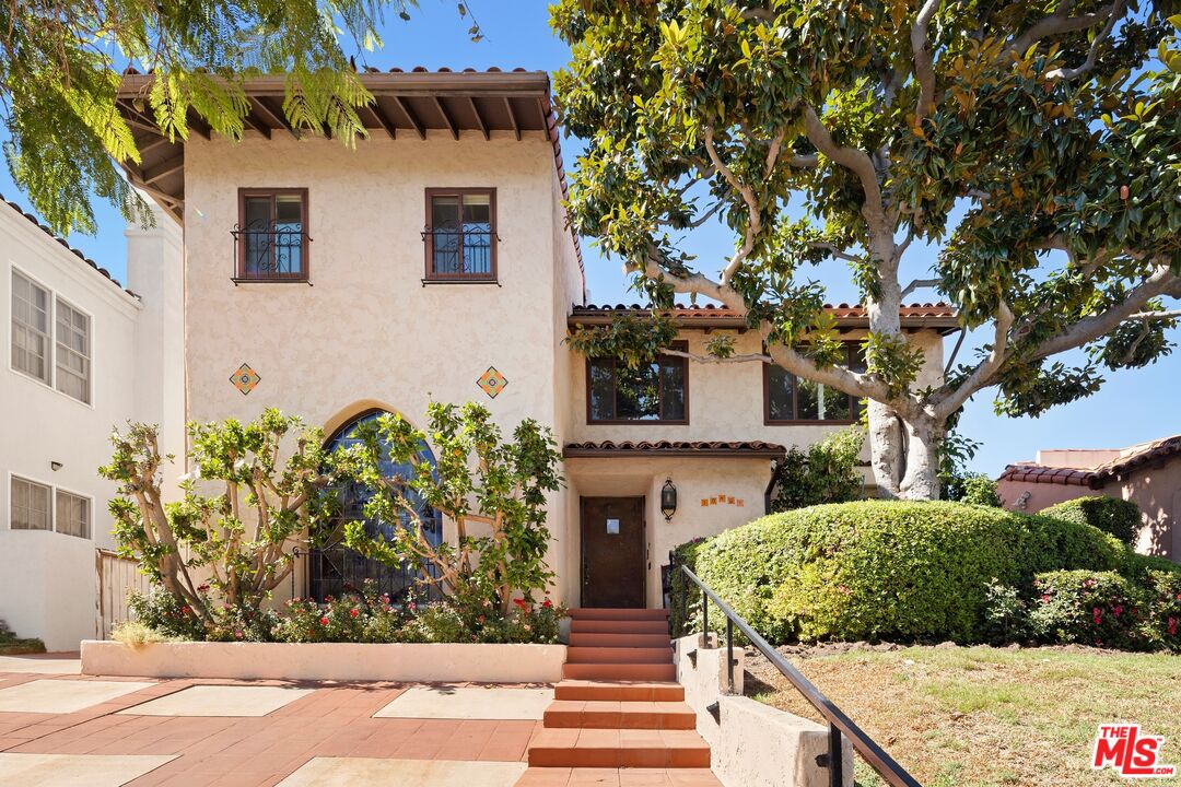 Two-story Spanish-style stucco home with an arched entry, brick steps, and a large shade tree.