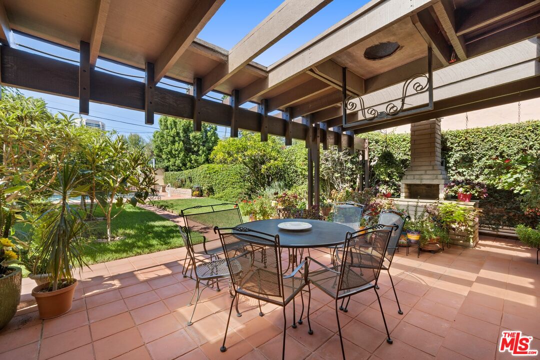 Outdoor dining patio beneath a wooden pergola, overlooking a landscaped garden and pool.