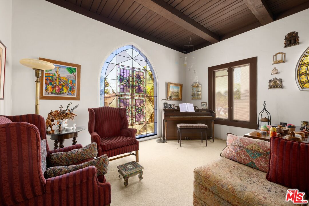 Living room with an arched stained-glass window, exposed wood ceiling beams, piano, and red armchairs.