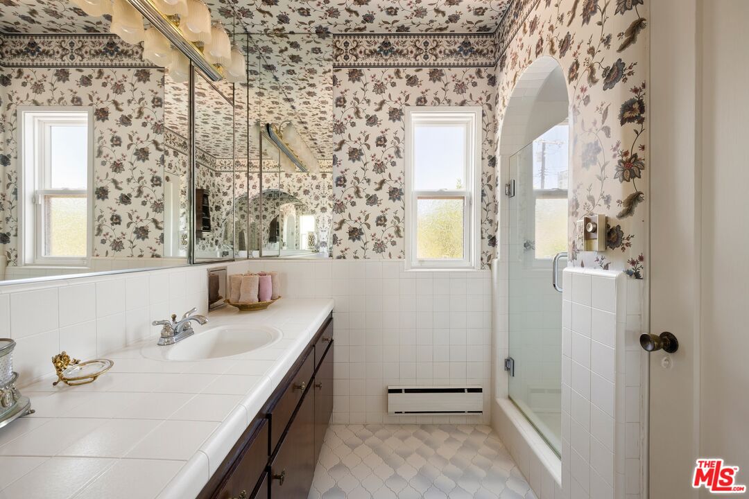 Bathroom with a long tiled vanity, mirrored wall, floral wallpaper, and glass-enclosed tub or shower.