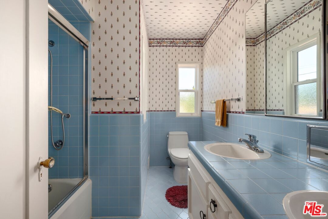 Bathroom with blue tile, patterned wallpaper, a large mirror, and a tub-shower.