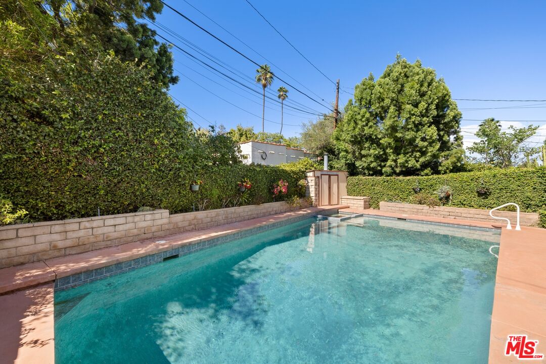 Backyard swimming pool with a pink stone deck and tall privacy hedges.