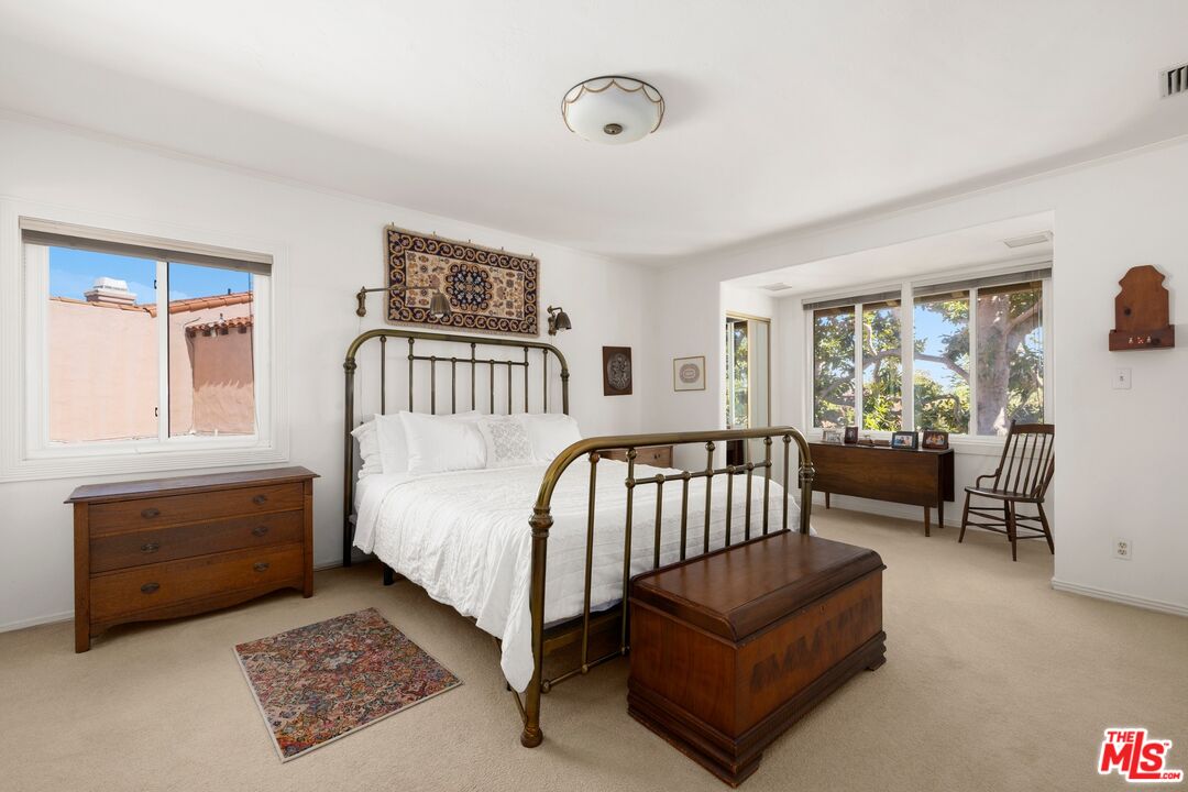 Furnished bedroom with a brass bed frame, wooden chest, and windows overlooking trees.