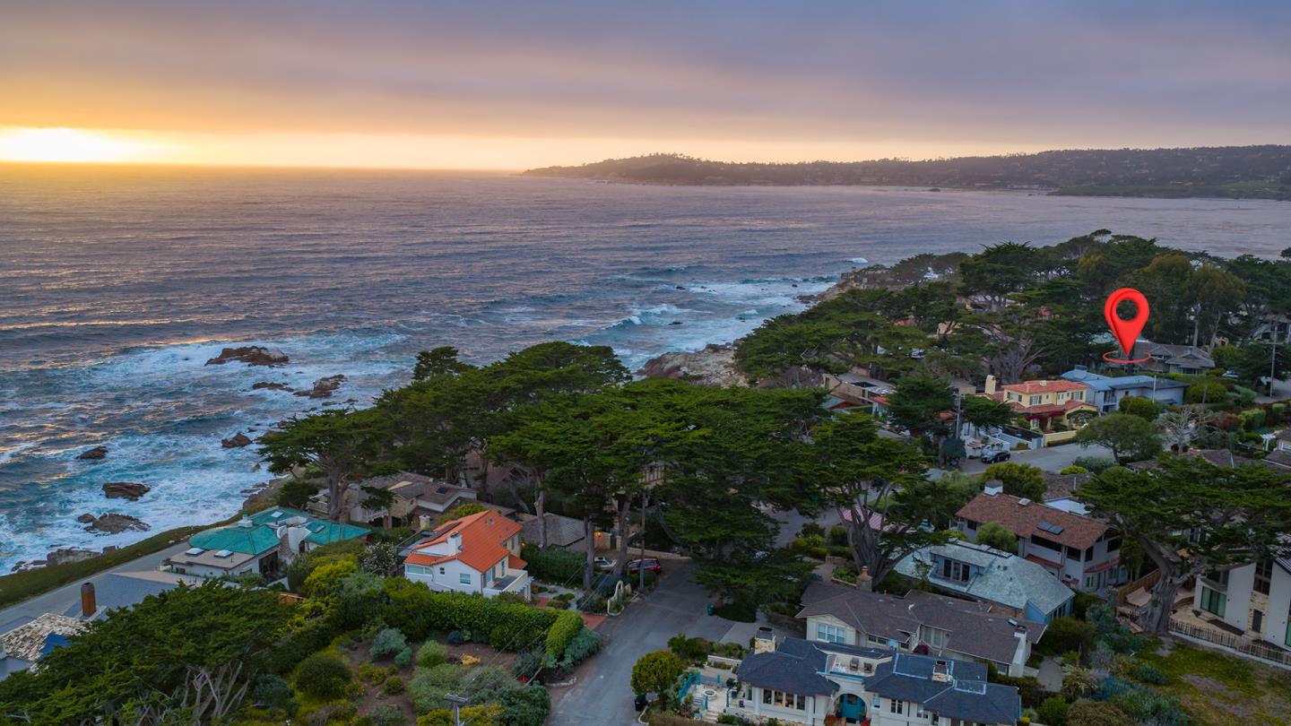 Aerial coastal neighborhood at sunset, with ocean and a red marker over one home.
