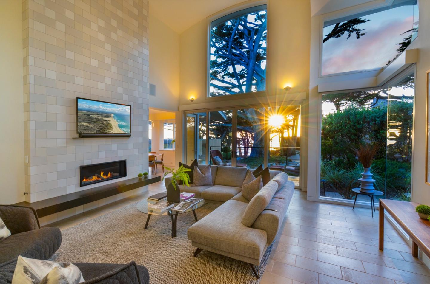 Furnished living room with a tiled fireplace wall, vaulted ceiling, and large windows facing trees.