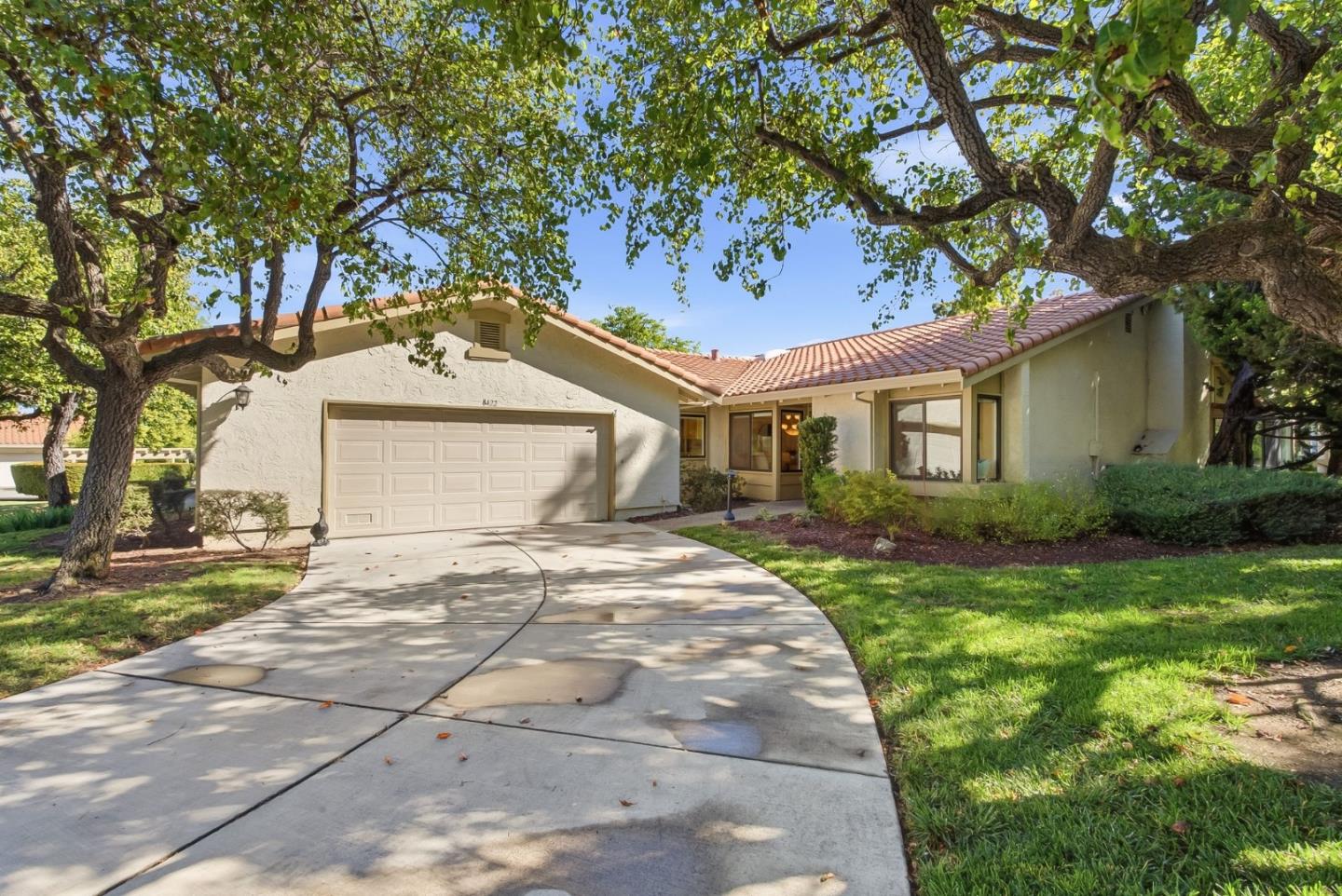 Single-story stucco home with a red tile roof, two-car garage, and curved driveway beneath mature trees.