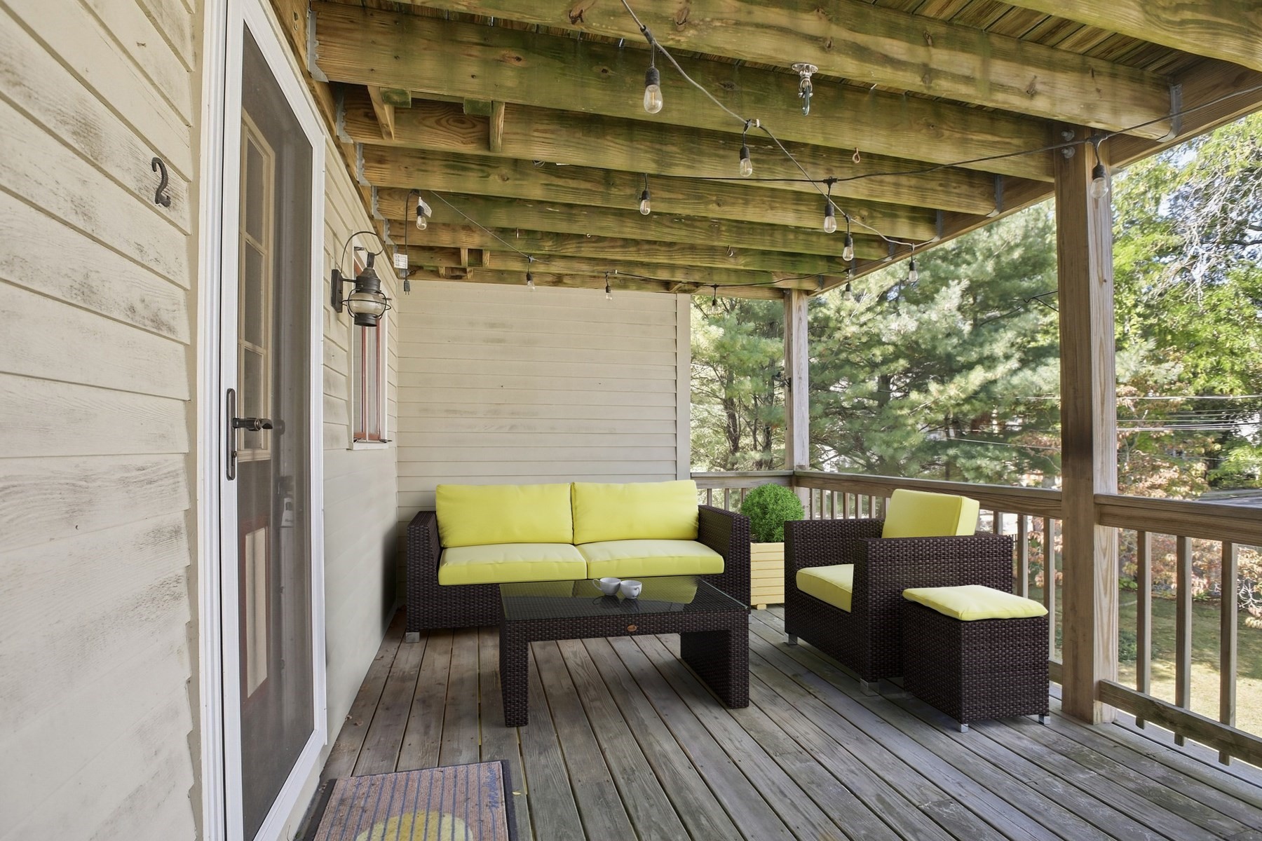 Covered wooden porch with wicker seating, bright cushions, and leafy trees beyond the railing.