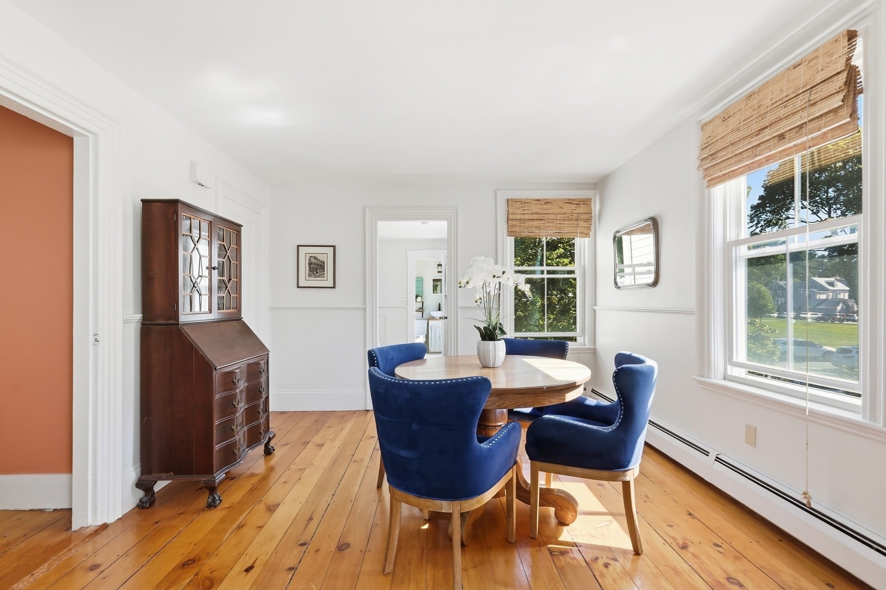 Dining room with a round table, blue chairs, polished wood floors, and large shaded windows.