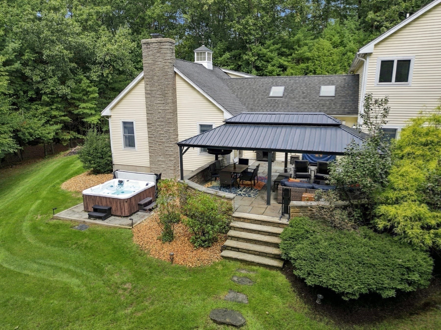 Rear of a beige home with a covered stone patio, outdoor seating, hot tub, and wooded lawn.