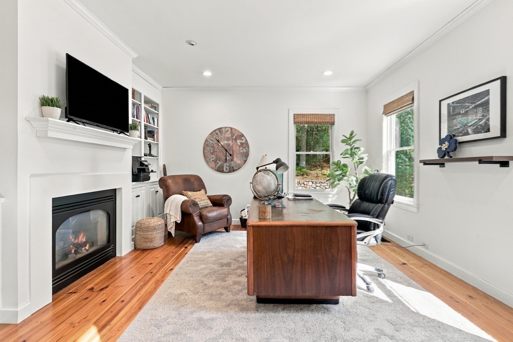 Home office with a wooden desk, leather chairs, and a fireplace beneath a mounted television.